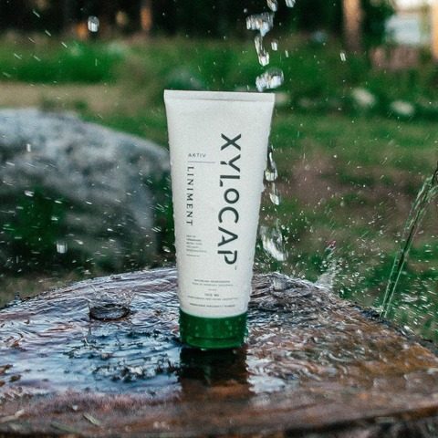 Xylocap Aktiv värmande liniment i tub med grön kork, stående på en våt trästubbe i skogsmiljö med vattenstänk